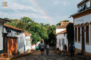 Uma rua movimentada e ensolarada em uma cidade brasileira, com calçadas largas. À esquerda, há uma padaria com uma porta de vidro e um toldo listrado. À direita, um banco com uma fachada moderna e um caixa eletrônico visível. No meio da rua, um ônibus amarelo está parado em um ponto. Mais à frente, pode-se ver uma praça com árvores e um banco de jardim.