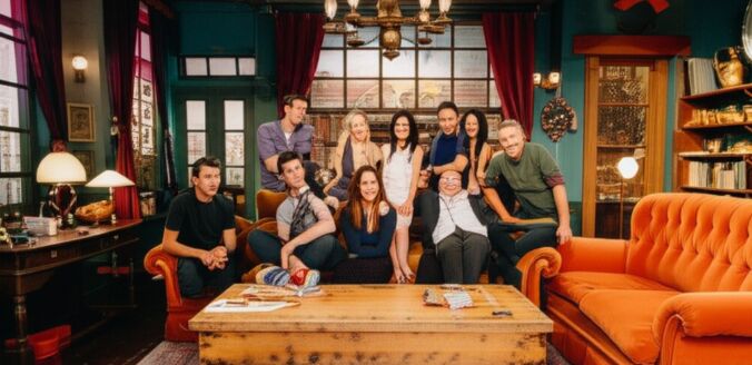 Seis amigos da série Friends (Chandler, Monica, Rachel, Joey, Phoebe e Ross) sorriem enquanto estão sentados e em pé atrás do famoso sofá laranja do Central Perk, em uma cena interna do café com iluminação quente, mesas de madeira e uma grande janela ao fundo.